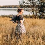 A young woman stands in a sunlit field by a lake, holding lavender flowers and smiling.