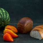 Loaves of bread with a watermelon and peppers arranged on a dark background, highlighting fresh produce.